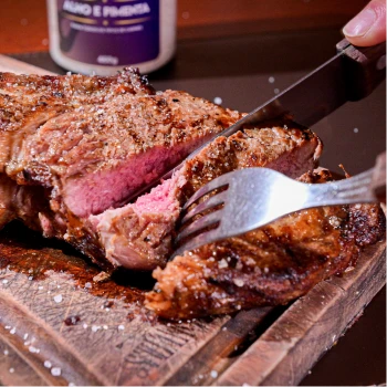 Close-up de um suculento bife ancho mal passado sendo cortado com garfo e faca em uma tábua de madeira.