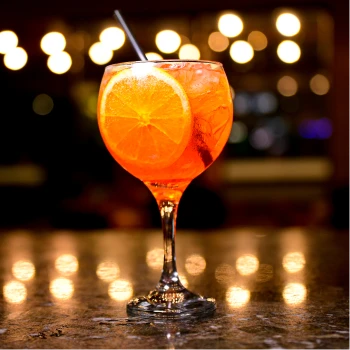 Taça de drink Aperol Spritz com uma fatia de laranja, servido no bar do Steak Ranch com luzes ao fundo.