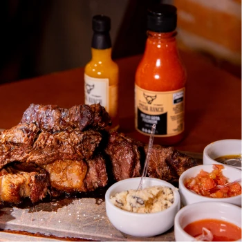 Porção de picanha fatiada servida na pedra com potes de molho vinagrete, farofa e chimichurri.