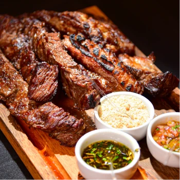 Tábua de carnes com diversos cortes de carne grelhada e fatiada, acompanhada por farofa e molhos.