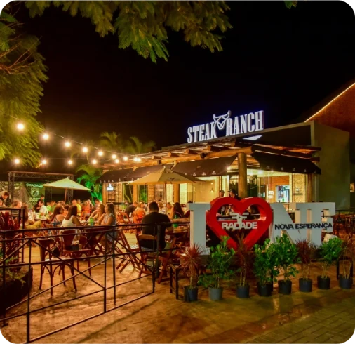 Vista noturna da área externa do restaurante Steak Ranch em Maringá, iluminada por luzes de cordão, com clientes nas mesas e a placa luminosa 'STEAK RANCH' visível.